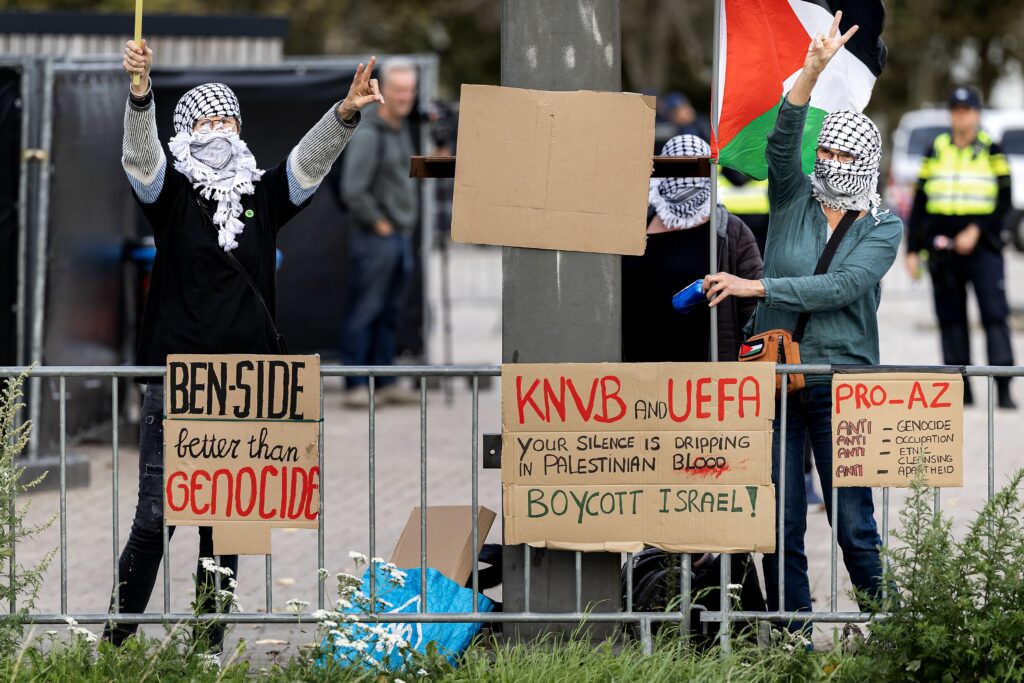 Protesters met gezichtsbedekking en Palestijnse vlag bij een afzetting, met borden tegen Israël en AZ.