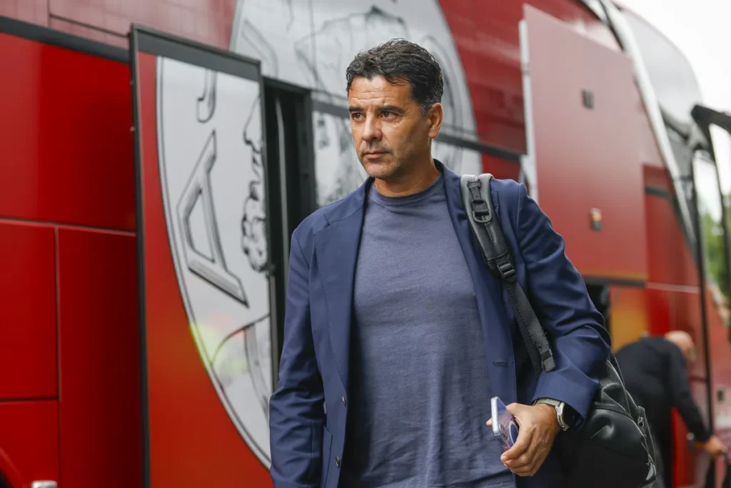 Míchel Sánchez loopt naast een rood teambusvoertuig met een tas over zijn schouder.