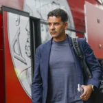 Míchel Sánchez loopt naast een rood teambusvoertuig met een tas over zijn schouder.