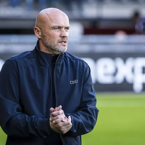 Een kale voetbaltrainer in een donker jack staat langs het veld en kijkt geconcentreerd toe.