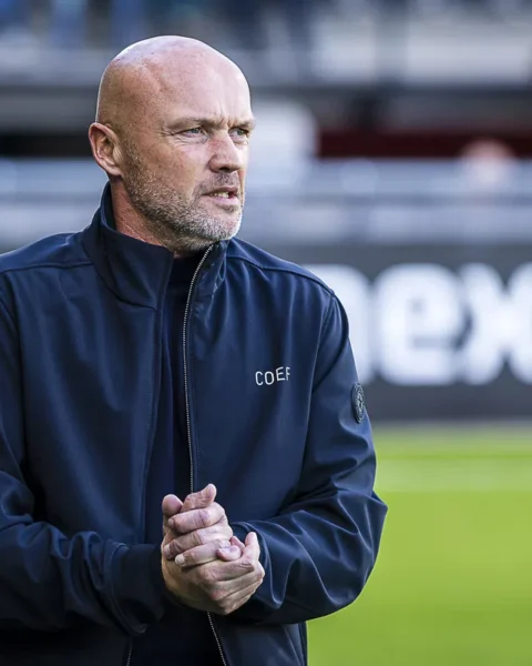 Een kale voetbaltrainer in een donker jack staat langs het veld en kijkt geconcentreerd toe.