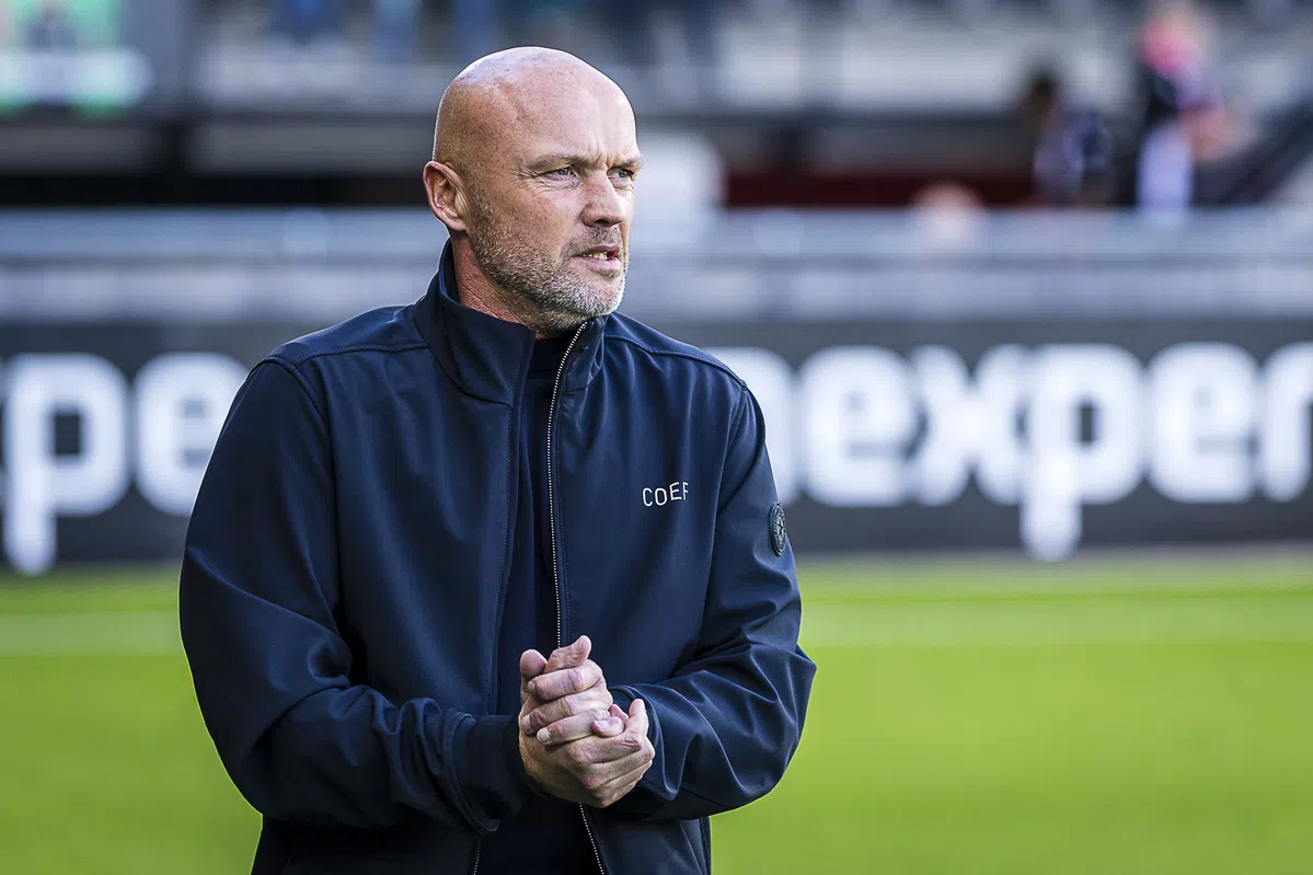 Een kale voetbaltrainer in een donker jack staat langs het veld en kijkt geconcentreerd toe.