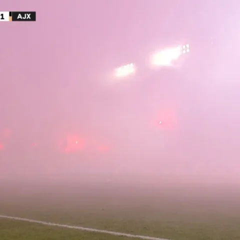 Een voetbalveld in dichte rook tijdens de wedstrijd Hajduk Split tegen Ajax, met de scorebug bovenin beeld.