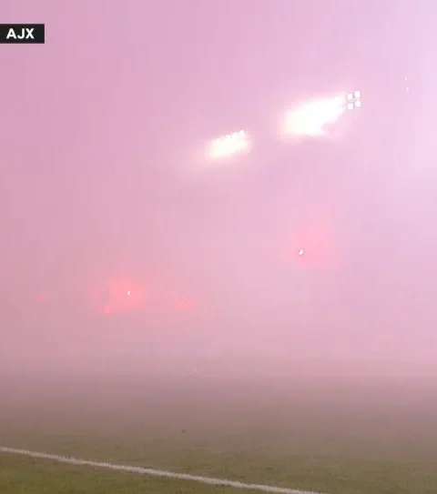 Een voetbalveld in dichte rook tijdens de wedstrijd Hajduk Split tegen Ajax, met de scorebug bovenin beeld.
