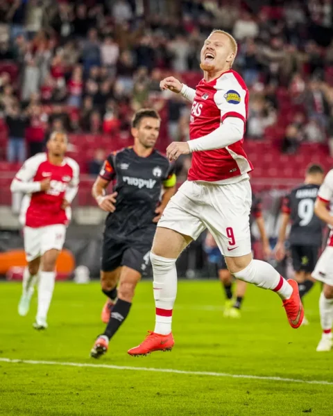 AZ-speler springt juichend op tijdens een wedstrijd, met tegenstanders op de achtergrond en de bal links op het veld.