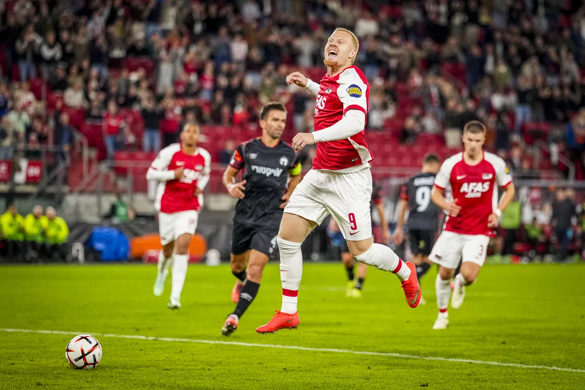 AZ-speler springt juichend op tijdens een wedstrijd, met tegenstanders op de achtergrond en de bal links op het veld.