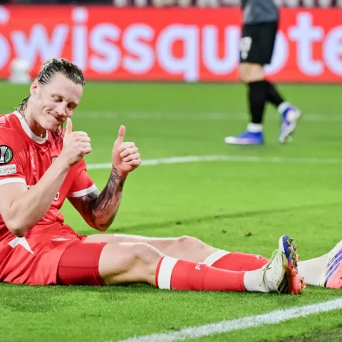 Voetballer in rood tenue zit glimlachend op het veld en steekt twee duimen op.