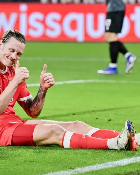 Voetballer in rood tenue zit glimlachend op het veld en steekt twee duimen op.