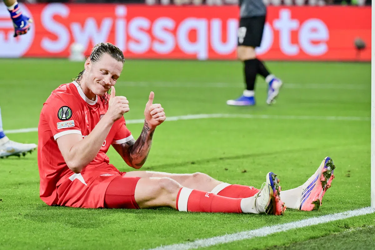 Voetballer in rood tenue zit glimlachend op het veld en steekt twee duimen op.