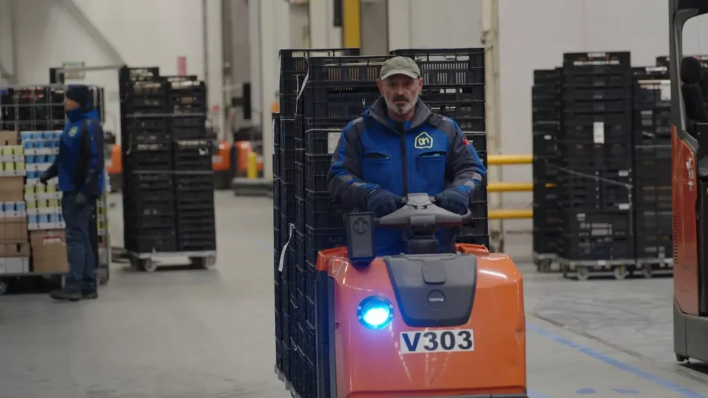 Medewerker rijdt met een elektrische magazijntruck tussen stellingen en rolcontainers in een distributiehal.