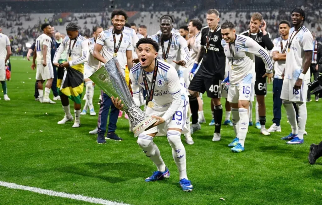 Jadon Sancho viert een overwinning met een trofee op het veld, omringd door ploeggenoten.