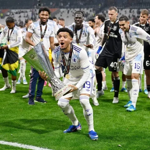 Jadon Sancho viert een overwinning met een trofee op het veld, omringd door ploeggenoten.