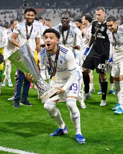 Jadon Sancho viert een overwinning met een trofee op het veld, omringd door ploeggenoten.