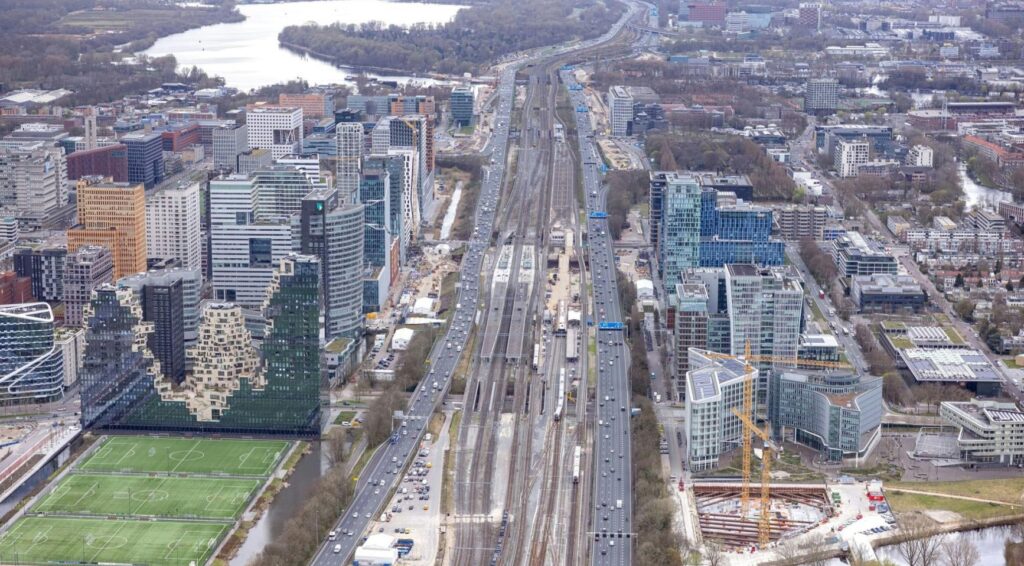 Luchtfoto van een stad met brede snelwegen, spoorlijnen en hoogbouw, met bouwwerkzaamheden langs de infrastructuur.