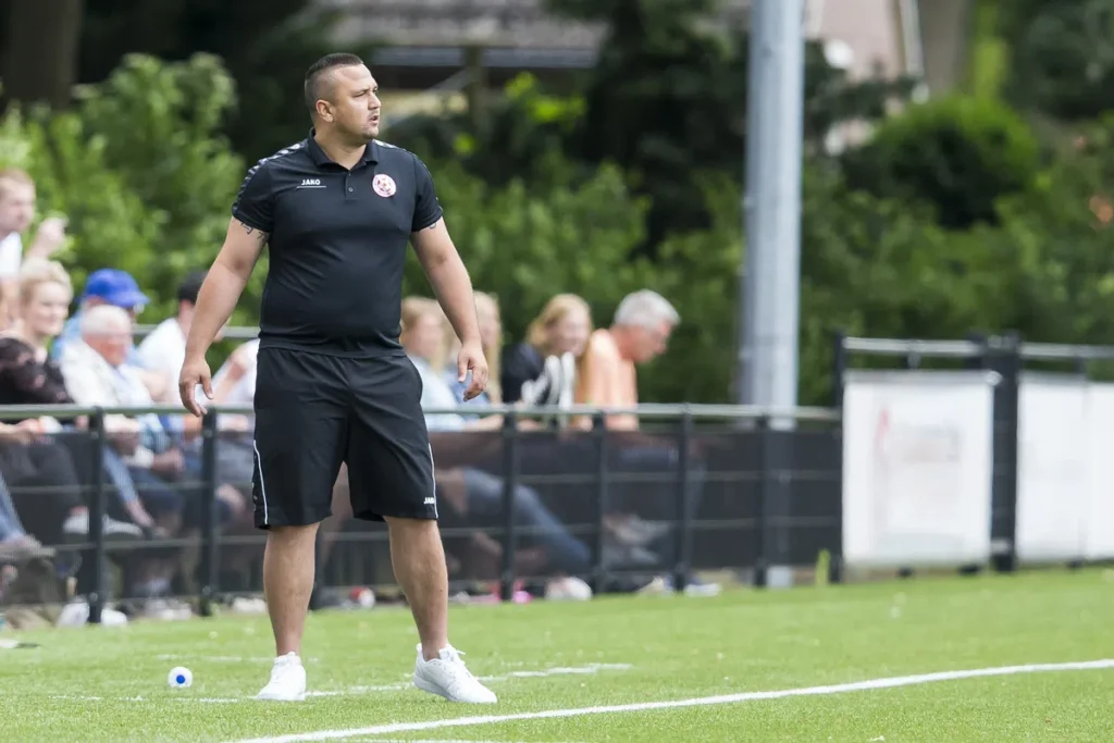 Jhon van Beukering staat langs de zijlijn op een voetbalveld in zwarte sportkleding.