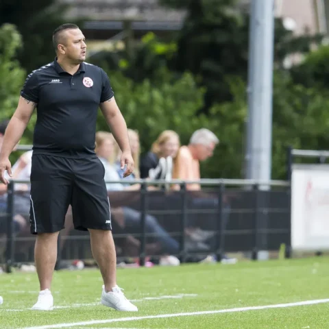 Jhon van Beukering staat langs de zijlijn op een voetbalveld in zwarte sportkleding.