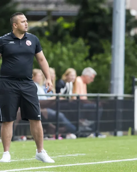 Jhon van Beukering staat langs de zijlijn op een voetbalveld in zwarte sportkleding.
