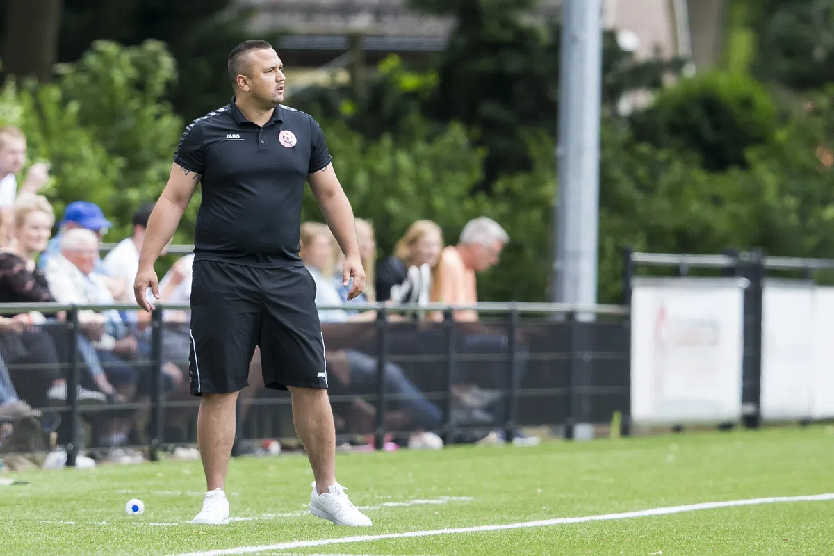 Jhon van Beukering staat langs de zijlijn op een voetbalveld in zwarte sportkleding.