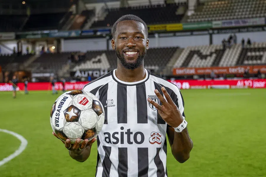 Een lachende Heracles Almelo-speler poseert op het veld met een voetbal.