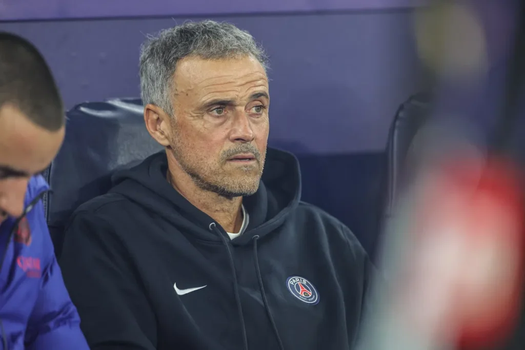 Luis Enrique zit op de bank in een PSG-trui en kijkt geconcentreerd vooruit.