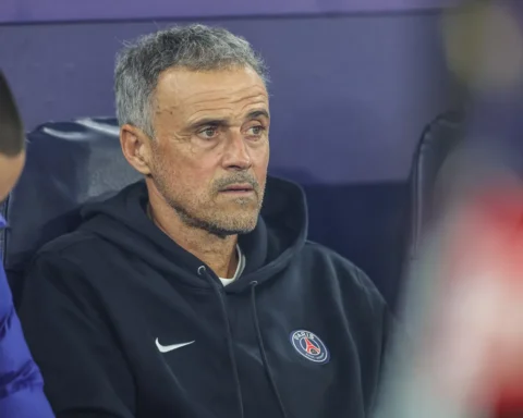 Luis Enrique zit op de bank in een PSG-trui en kijkt geconcentreerd vooruit.