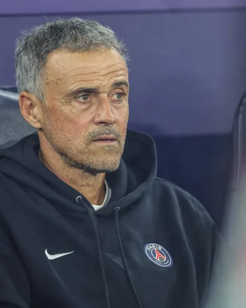 Luis Enrique zit op de bank in een PSG-trui en kijkt geconcentreerd vooruit.