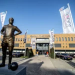 Standbeeld voor het Koning Willem II-stadion met clubvlaggen en geparkeerde auto’s.