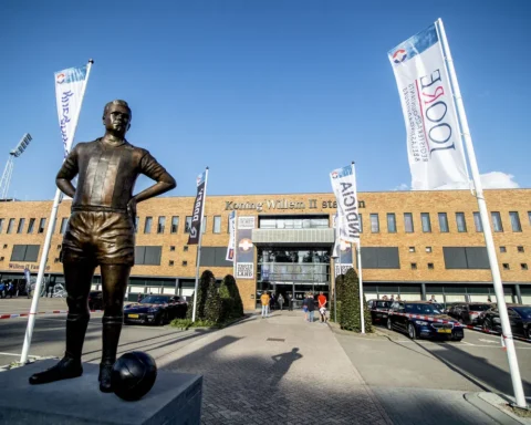 Standbeeld voor het Koning Willem II-stadion met clubvlaggen en geparkeerde auto’s.