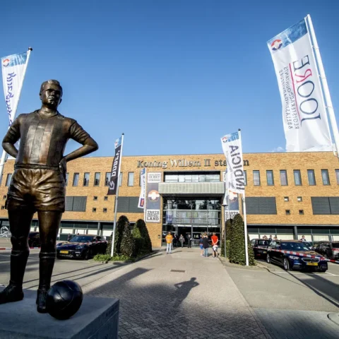 Standbeeld voor het Koning Willem II-stadion met clubvlaggen en geparkeerde auto’s.