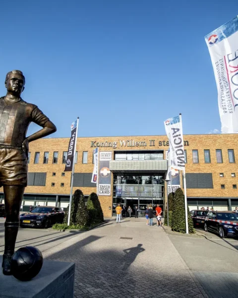 Standbeeld voor het Koning Willem II-stadion met clubvlaggen en geparkeerde auto’s.