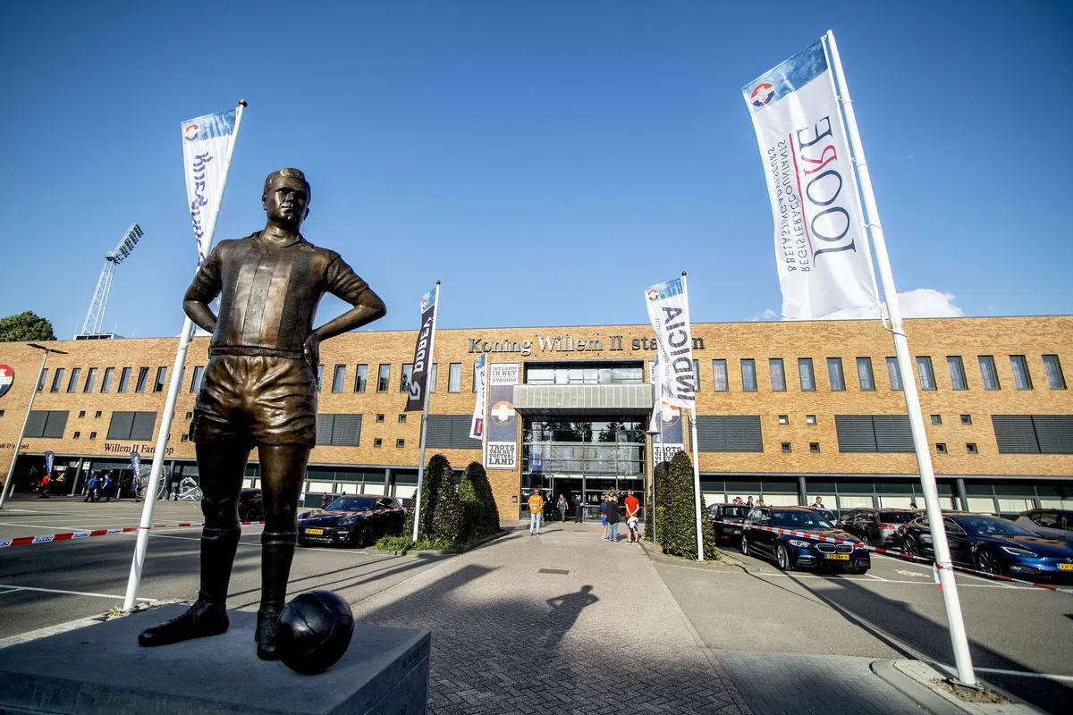 Standbeeld voor het Koning Willem II-stadion met clubvlaggen en geparkeerde auto’s.