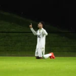 Een voetballer in wit tenue knielt op het veld en maakt een gebaar met zijn handen.