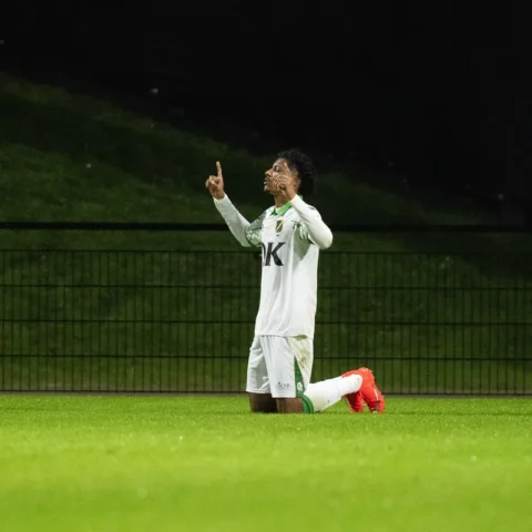 Een voetballer in wit tenue knielt op het veld en maakt een gebaar met zijn handen.