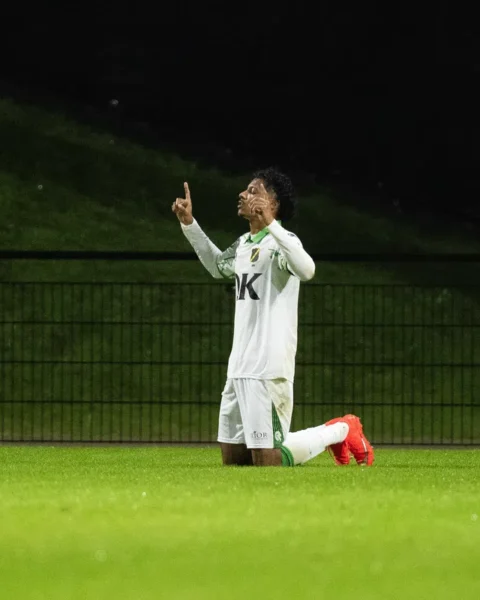 Een voetballer in wit tenue knielt op het veld en maakt een gebaar met zijn handen.