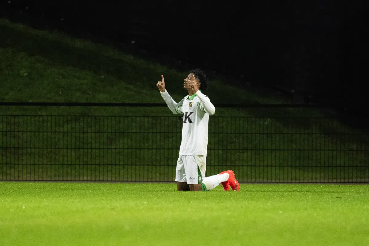 Een voetballer in wit tenue knielt op het veld en maakt een gebaar met zijn handen.