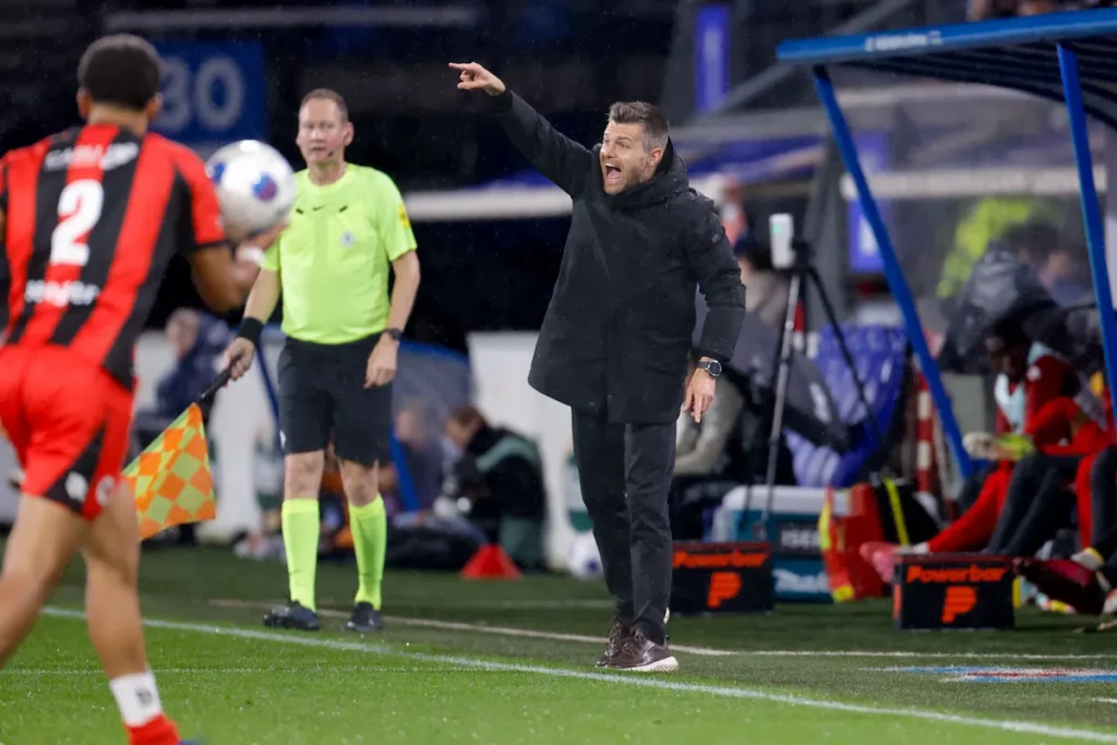 Trainer langs de lijn bij een voetbalwedstrijd, wijzend en roepend terwijl een grensrechter en speler in beeld zijn.