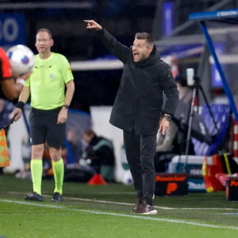 Trainer langs de lijn bij een voetbalwedstrijd, wijzend en roepend terwijl een grensrechter en speler in beeld zijn.