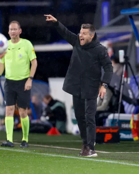 Trainer langs de lijn bij een voetbalwedstrijd, wijzend en roepend terwijl een grensrechter en speler in beeld zijn.
