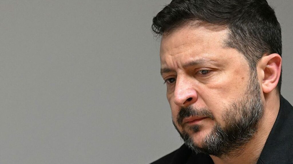 Portret van Volodymyr Zelensky met ernstige blik tegen een neutrale achtergrond.
