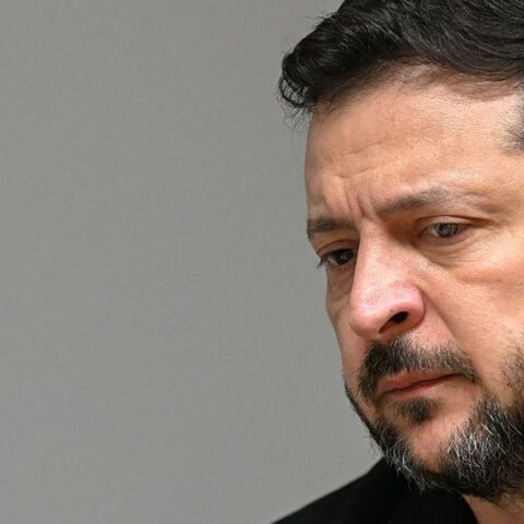 Portret van Volodymyr Zelensky met ernstige blik tegen een neutrale achtergrond.