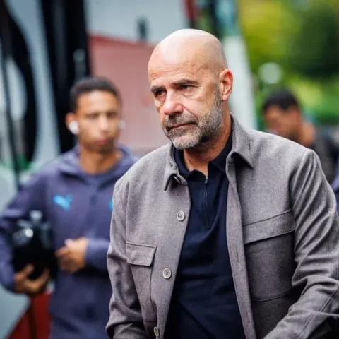 Peter Bosz loopt buiten bij een voetbalomgeving, met staff op de achtergrond.