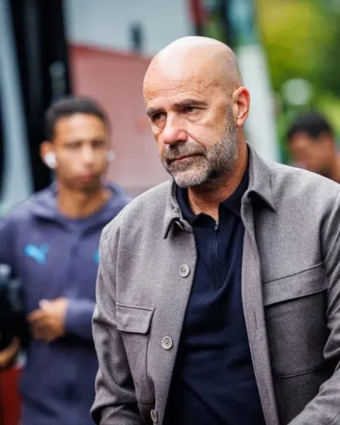 Peter Bosz loopt buiten bij een voetbalomgeving, met staff op de achtergrond.