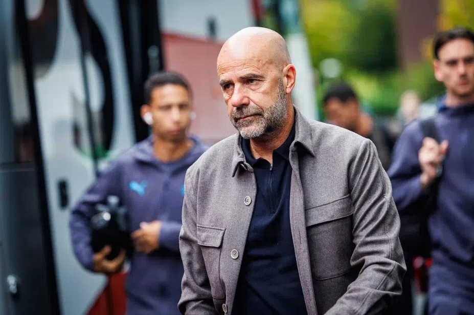 Peter Bosz loopt buiten bij een voetbalomgeving, met staff op de achtergrond.