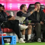 Vincent Kompany sits on the Bayern München bench during a match, with staff and branded seating around him.