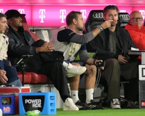 Vincent Kompany sits on the Bayern München bench during a match, with staff and branded seating around him.