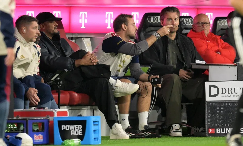 Vincent Kompany sits on the Bayern München bench during a match, with staff and branded seating around him.