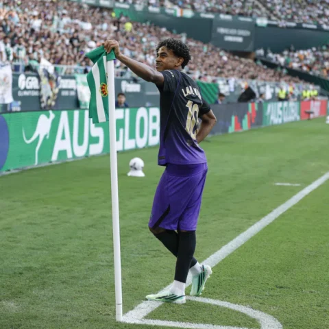 Lamine Yamal staat bij de hoekvlag in een stadion, met een groen-witte vlag in zijn hand.
