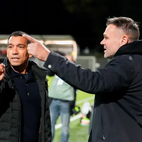 Giovanni van Bronckhorst staat langs de lijn en praat met een andere man tijdens een voetbalwedstrijd.