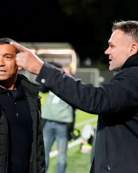 Giovanni van Bronckhorst staat langs de lijn en praat met een andere man tijdens een voetbalwedstrijd.
