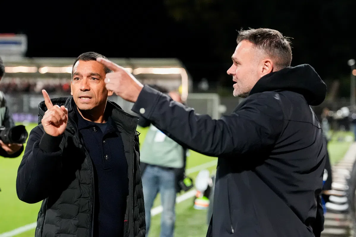 Giovanni van Bronckhorst staat langs de lijn en praat met een andere man tijdens een voetbalwedstrijd.